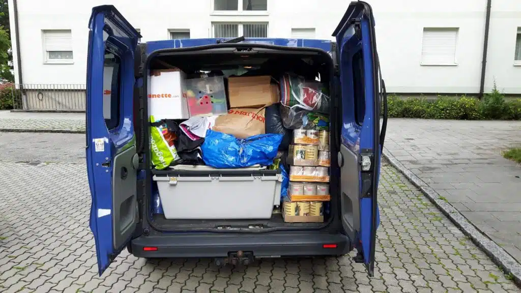 Ein geöffneter blauer Transporter mit Säcken und Kartons steht auf Pflaster vor einem hellen Gebäude